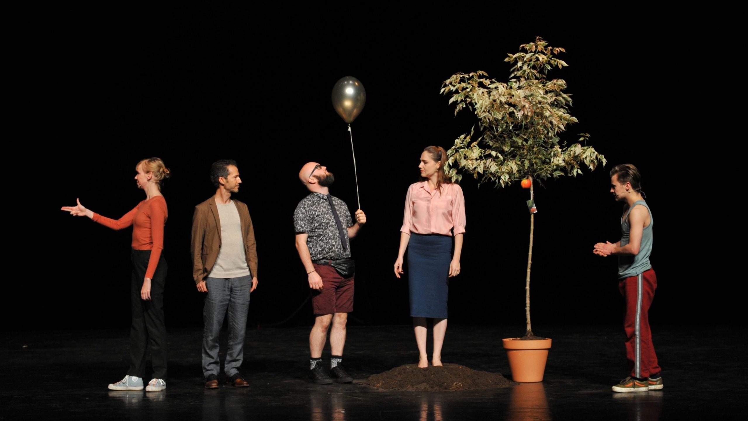 Fem personer står på scen bredvid en träd. alla tittar åt olika håll och en man håller en ballong.